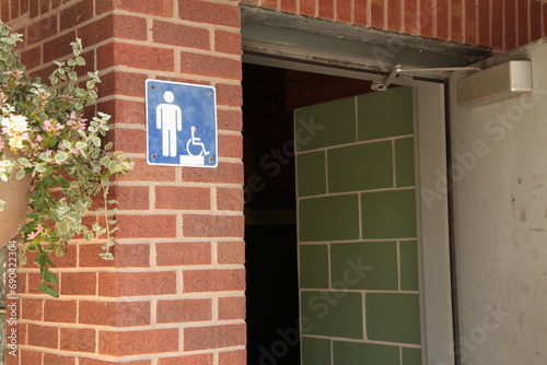 male bathroom washroom mens room square blue white symbol sign and handicap on brick wall outside outdoors exterior next to open gray bathroom door and branches leaves