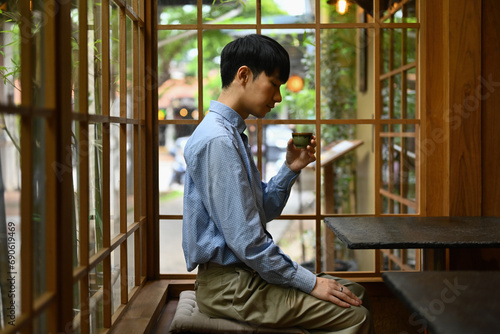 A young Asian man sips a small cup of hot tea in a quiet, traditional Japanese-style restaurant or cafe