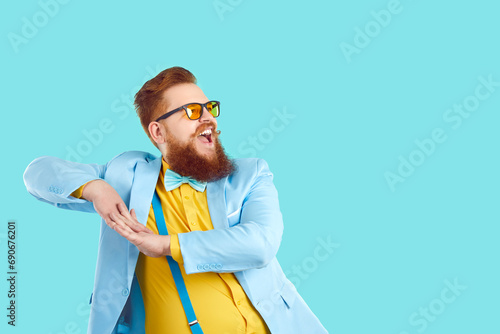Funny confident fat man in trendy modern outfit having fun at party. Happy overweight bearded guy in cool spectacles, blue suit, yellow shirt, and suspenders dancing on turquoise copy space background