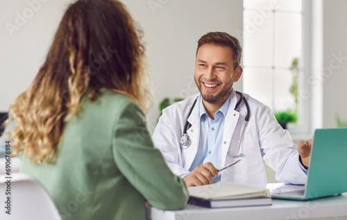 Cheerful friendly male doctor at medical office in hospital, showing examination results on laptop monitor screen and having consultation with young woman patient sitting in clinic.
