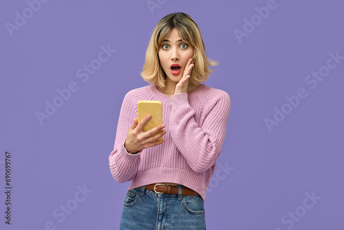 Shocked young gen z blonde woman shopper holding smartphone using mobile cell phone surprised about wow fashion sale app, omg promo discounts on cellphone isolated on purple background.