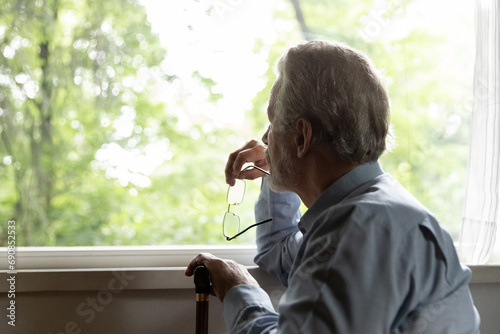 Retirement problem. Pensive old man nursing home patient sit by window hold glasses walking stick lost in sad thoughts. Upset aged male think about bad health loneliness miss deceased wife. Copy space