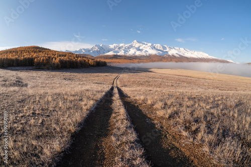 Remote dirt trail through the frozen steppe to snow mountains. Bright sunny autumn atmospheric minimalist landscape with a dirt path among the grasses in the highlands.