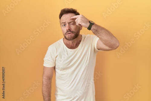 Middle age man with beard standing over yellow background pointing unhappy to pimple on forehead, ugly infection of blackhead. acne and skin problem