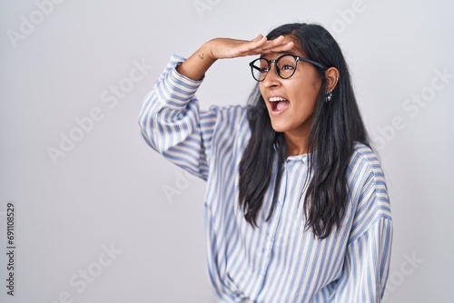 Young hispanic woman wearing glasses very happy and smiling looking far away with hand over head. searching concept.