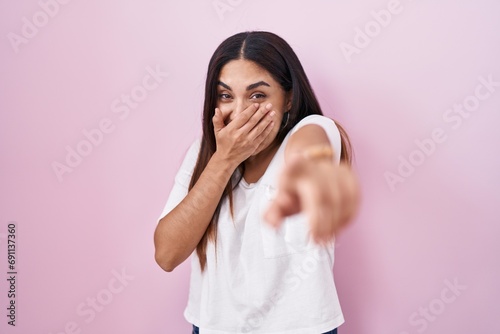 Young arab woman standing over pink background laughing at you, pointing finger to the camera with hand over mouth, shame expression