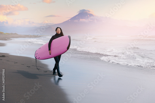 Extreme surfer happy young woman in wetsuit with surfboard go to winter surfing in Atlantic ocean Kamchatka Russia on background volcano