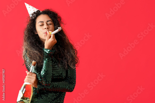 Beautiful young African-American woman with party whistle and bottle of champagne celebrating Birthday on red background