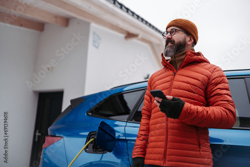 Man charging electric car during cold snowy day, using electric vehicle charging app, checking battery life, energy consumption on smart phone. Charging and driving electric vehicles during winter