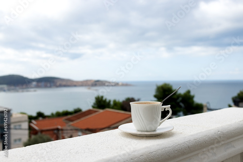 A white mug with coffee on a saucer stands on the balcony railing of the hotel room. Sea view from the apartment balcony