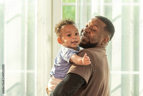 Happy african black parents dad father throw baby son on piggyback and neck riding teasing fun near window. Black baby son and daddy enjoy teasing kiss cheek and neck riding at window light