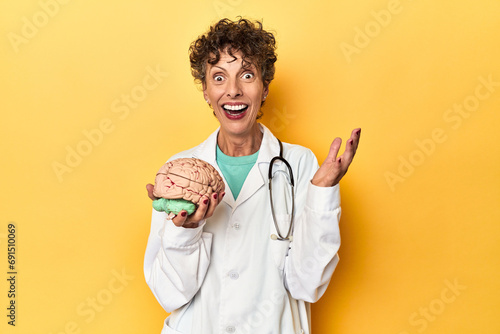 Doctor holding a brain model on yellow studio receiving a pleasant surprise, excited and raising hands.