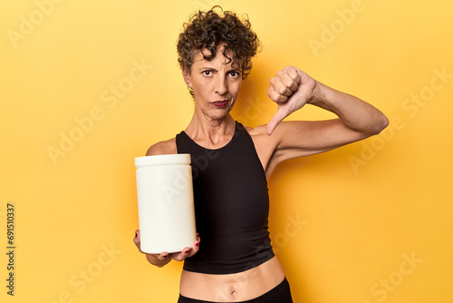 MIddle aged athlete woman holding protein supplement on yellow showing a dislike gesture, thumbs down. Disagreement concept.