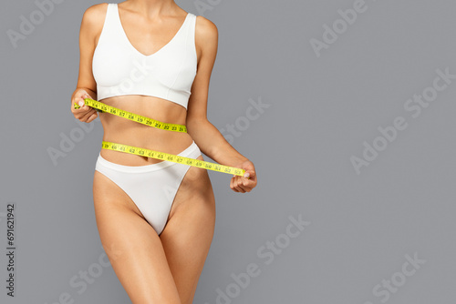 European woman in white sports bra and panties holds yellow measuring tape around waist