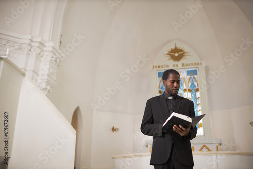 Wide angle portrait of African American man as priest reading Bible at church altar, copy space