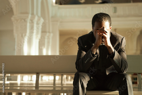Front view at young African American priest praying in church and holding rosary beads in sunlight, copy space