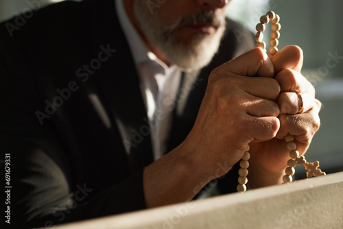 Close up of senior man holding rosary beads in prayer lit by sunlight rays, copy space