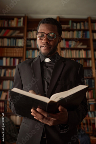 Vertical portrait of young African American man as priest reading Bible in church with books in background