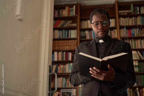 Waist up portrait of young Black priest reading Bible in office with bookcase in background, copy space