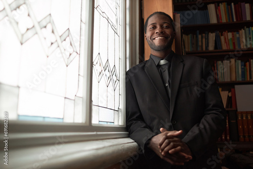 Waist up portrait of smiling African American priest looking at camera standing by window in church library, copy space