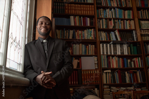 Portrait of smiling Black man as pastor looking at camera posing by window in library, copy space