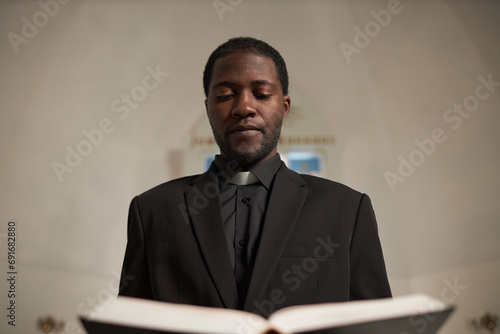 Waist up portrait of young Black priest reading Bible standing at altar during Sunday service