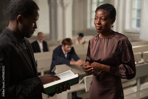 Waist up portrait of mature African American woman talking to priest during Sunday service in church