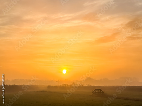 Bright orange sunrise at fields with light fog and a fence or gate in Holland