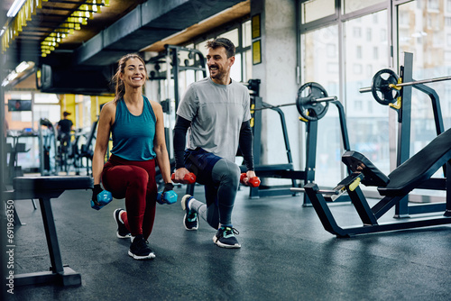 Happy athletic couple exercising with hand weights in lunge position in gym.