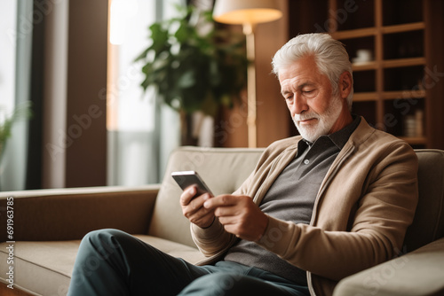 Relaxed and happy senior man with white grey hair using mobile phone sitting on sofa at home. Concept of technology and older people