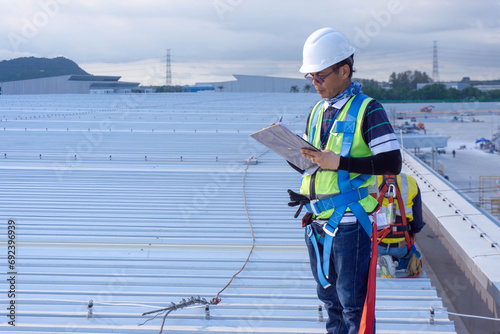 Engineer wearing full safety body harness working inspection defect work of metal roof sheet in site construction warehouse building
