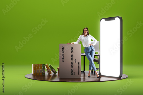 Smiling young woman standing with big credit card near giant 3D model of mobile phone with empty screen over green background. Online shopping