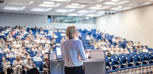 Female speaker giving a talk on corporate business conference. Unrecognizable people in audience at conference hall. Business and Entrepreneurship event