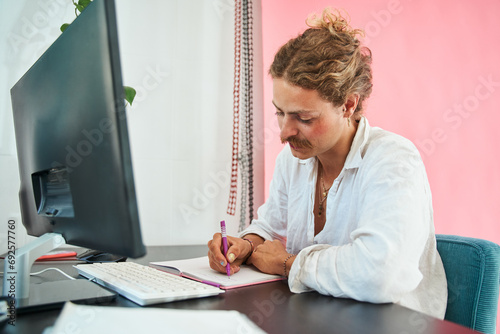 Creative young freelancer with face makeup sitting at computer table and writing notes