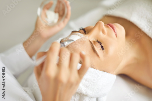 Woman getting professional facial treatment at spa and beauty salon. Beautician or cosmetologist holding brush and applying vitamin mask on face of young relaxed woman. Close up. Skin care concept