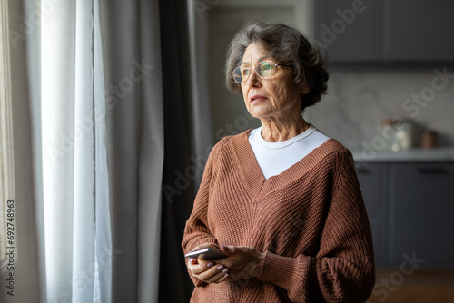 Worried depressed senior woman stand by window look away troubled, holding cellphone in hands, lady lost in sad heavy thoughts