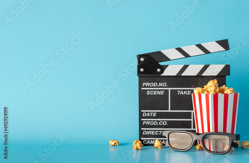 Experience excitement of much-awaited movie debut. Side view captures table scene with clapperboard, striped popcorn box, and 3D glasses against light blue wall, leaving space for text or film adverts