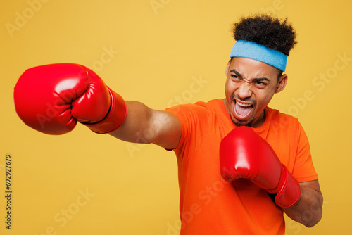 Young masculine fitness trainer instructor sporty man sportsman wear orange t-shirt boxing red gloves punch spend time in home gym isolated on plain yellow background. Workout sport fit abs concept.