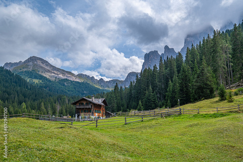 Nestled in Val di Funes, a wooden cabin stands against the Dolomites backdrop, surrounded by lush green pastures, a serene alpine retreat embraced by nature's tranquillity
