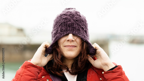 Portrait of a happy woman on the street covering her eyes with a wool hat in winter enjoying the freezing morning of the cold season. Nice woman playing with her winter hat on the street posing