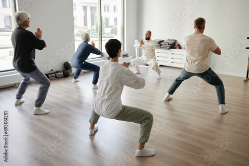 Back view at senior qigong students doing concentration exercise in half-squat position, male trainer showing prayer pose