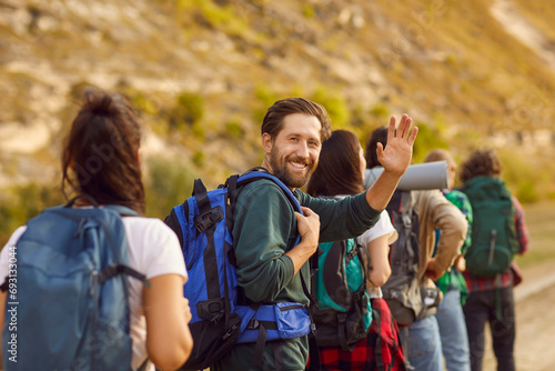 Portrait of a happy smiling bearded man waving hand looking cheerful at camera while trekking with backpack during a walking tour of the mountains with a group of his young friends.