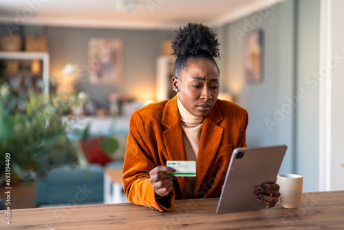 Worried young adult woman holding credit card while checking electronic bank account over digital tablet pc.