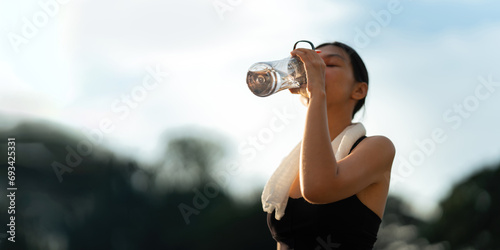 Beautiful young Asian woman stops to drinking water and wipe off her sweat after her evening run at park