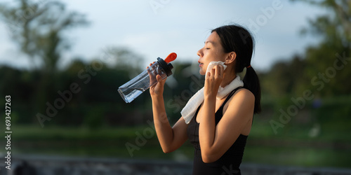 Beautiful young Asian woman stops to drinking water and wipe off her sweat after her evening run at park