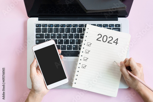 Flat lay of woman hand writing 2024 to do list on notebook while holding mobile phone with blank screen. Copy space, top view. 2024 planning concept