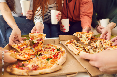 Close up hands of a young people friends gathering in pizzeria or at home together eating tasty Italian food taking pizza slices from box enjoying party with cardboard cups in hands.