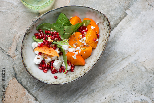 Bowl of fresh persimmon, goat cheese, spinach and pomegranate salad, above view on a grey granite background, horizontal shot with space