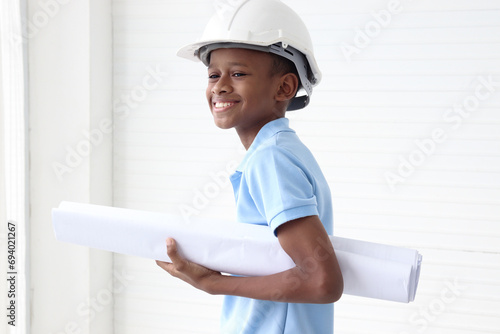 Happy smiling African boy wearing helmet safety hard hat, holding blueprint, standing on white wall room. Cute little child kid pretending to be an engineer.