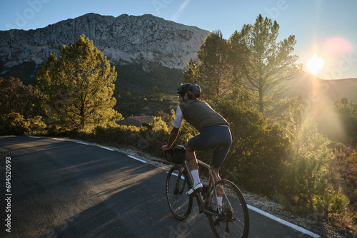 A man is riding a gravel bicycle on the road in the hills with a mountain view at sunset. Athlete wearing white and blue cycling kit and black helmet on a gravel bike. Cycling adventure. Bernia, Spain